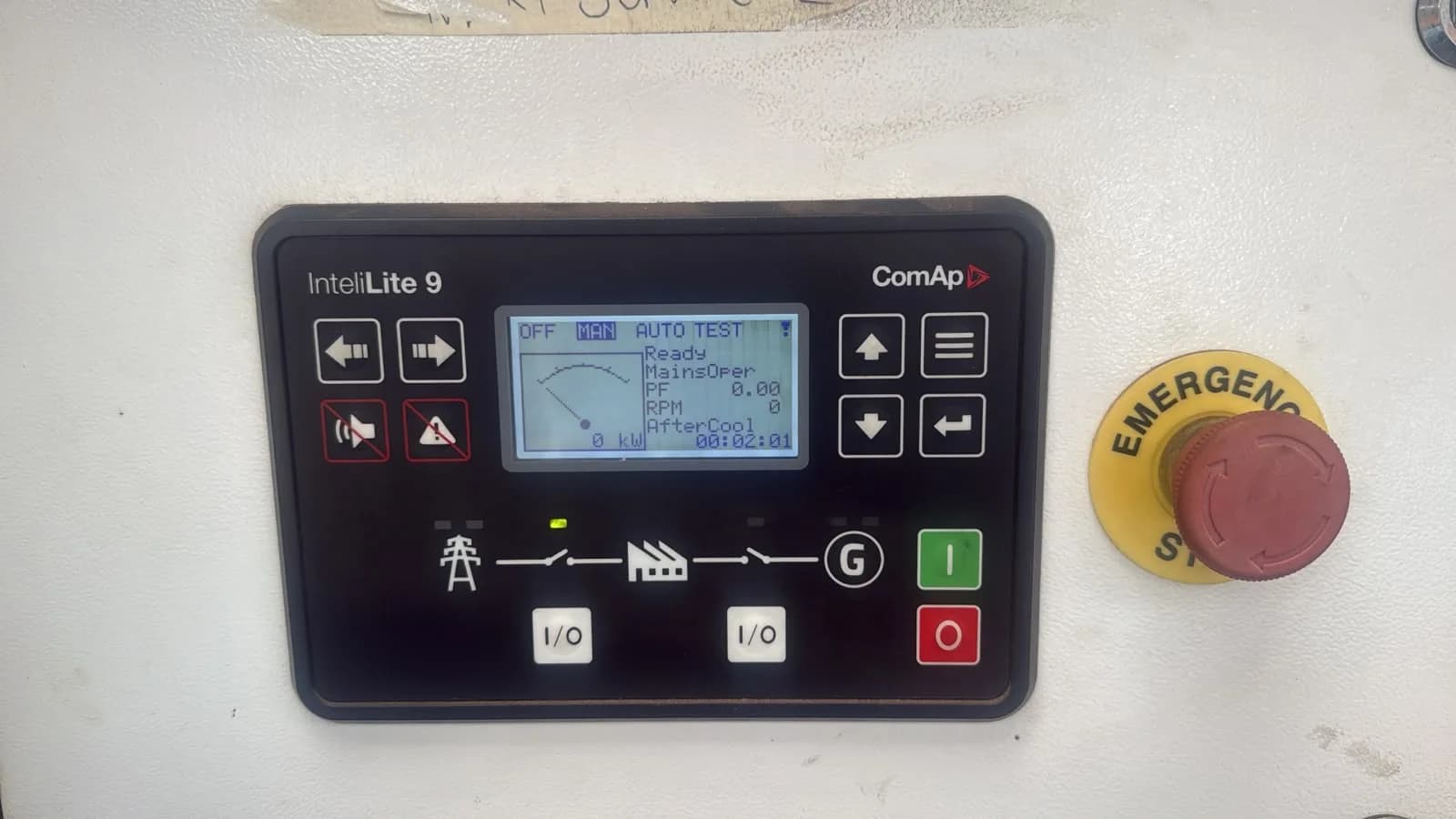 ComAp InteliLite 9 generator controller in manual mode beside a red emergency stop button on a cream panel