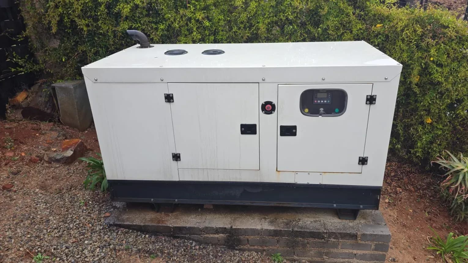White canopied diesel standby generator with controller window on a brick plinth among gravel and garden shrubs