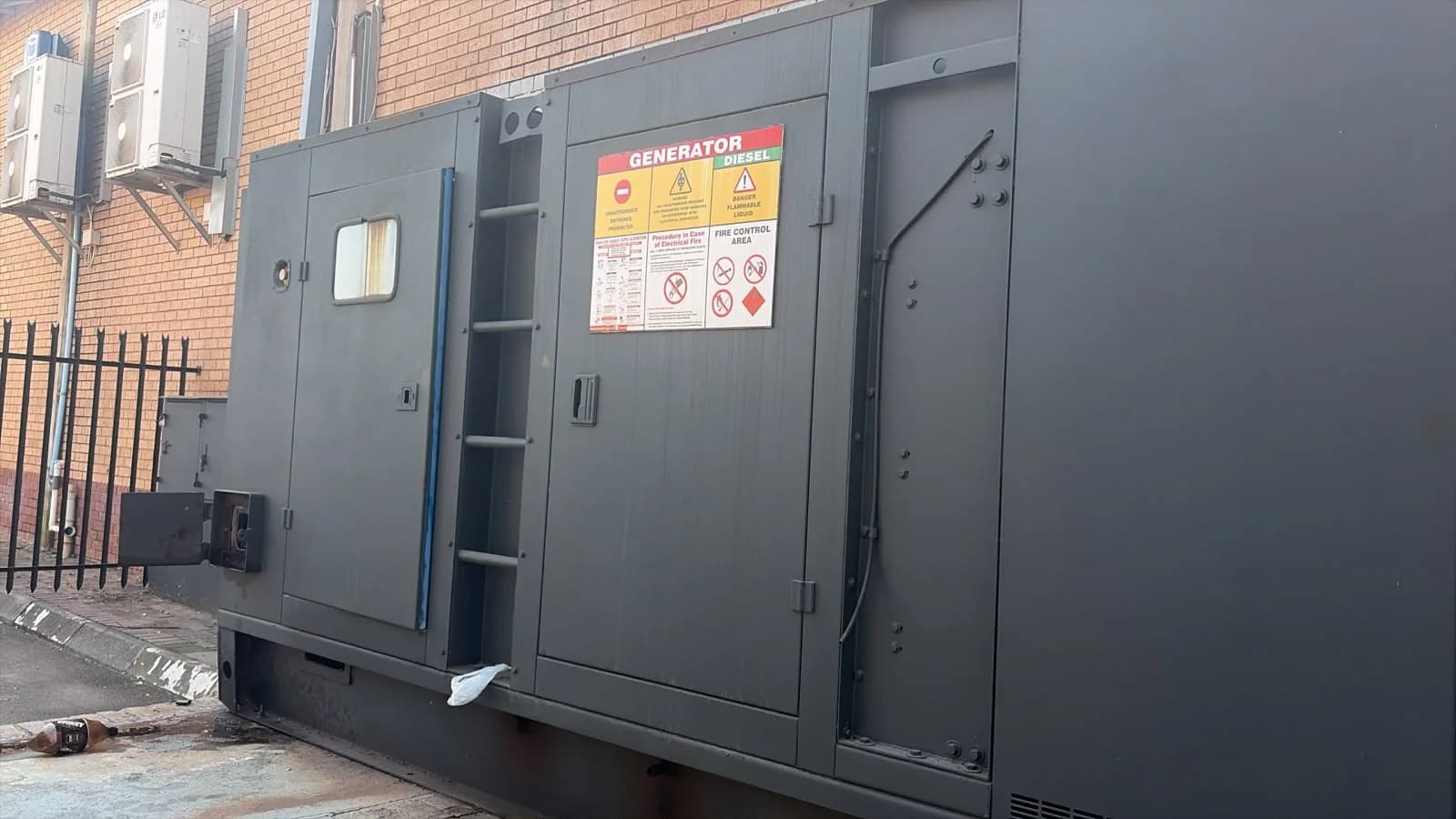 Large dark grey canopied diesel generator with built-in ladder and generator safety signs beside a face-brick building