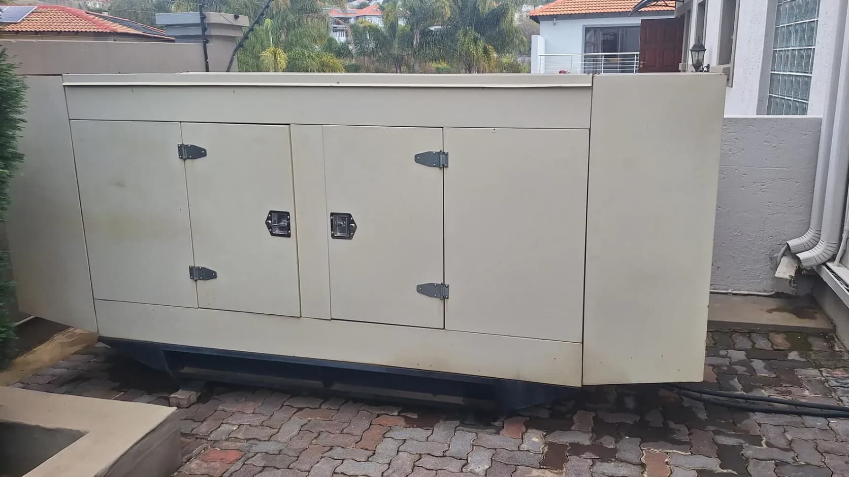 Large cream canopied diesel generator on brick paving beside a house, tiled roofs and palm trees behind