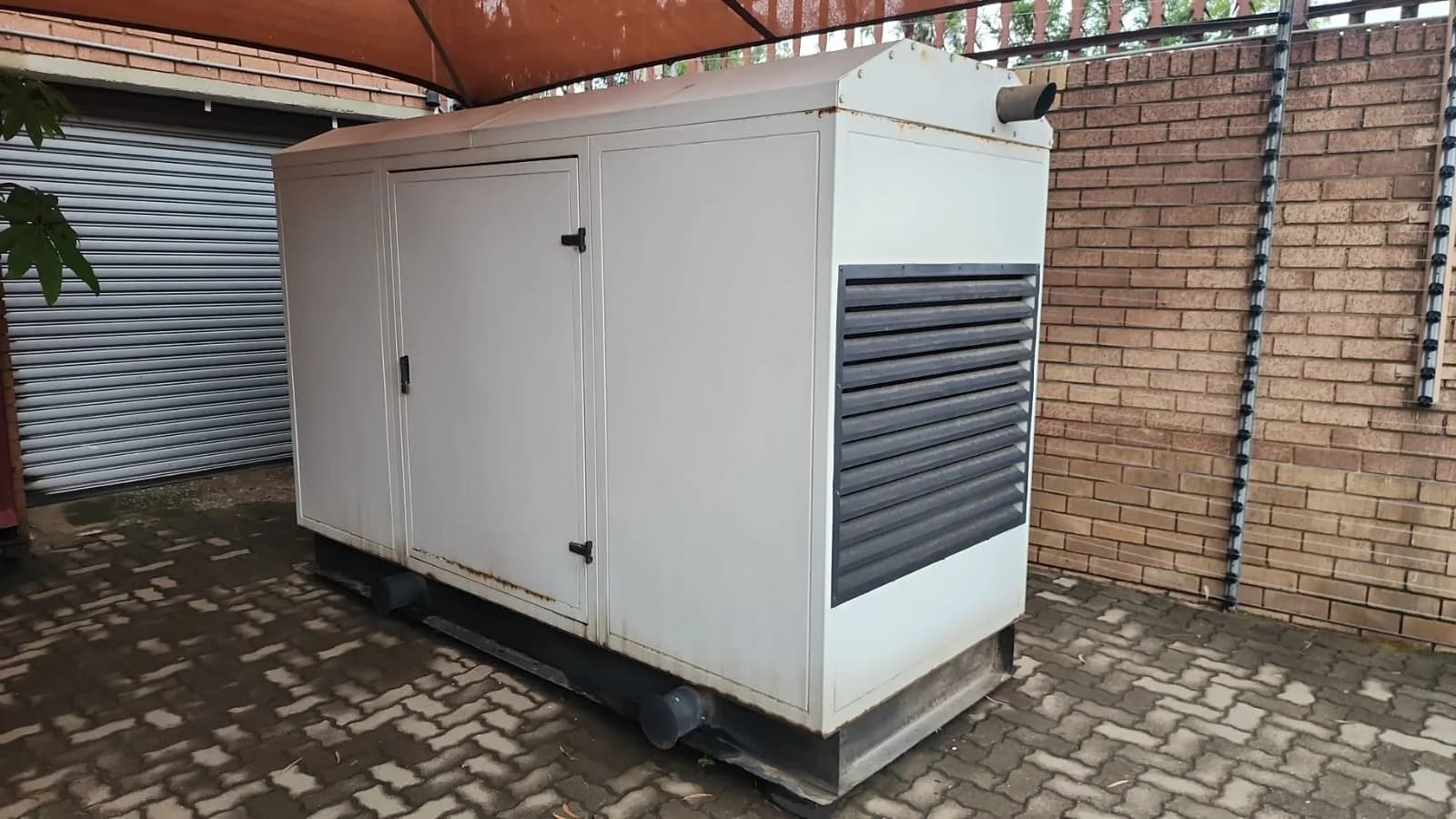 White canopied diesel generator with louvred radiator end on brick paving under a carport beside a face-brick wall