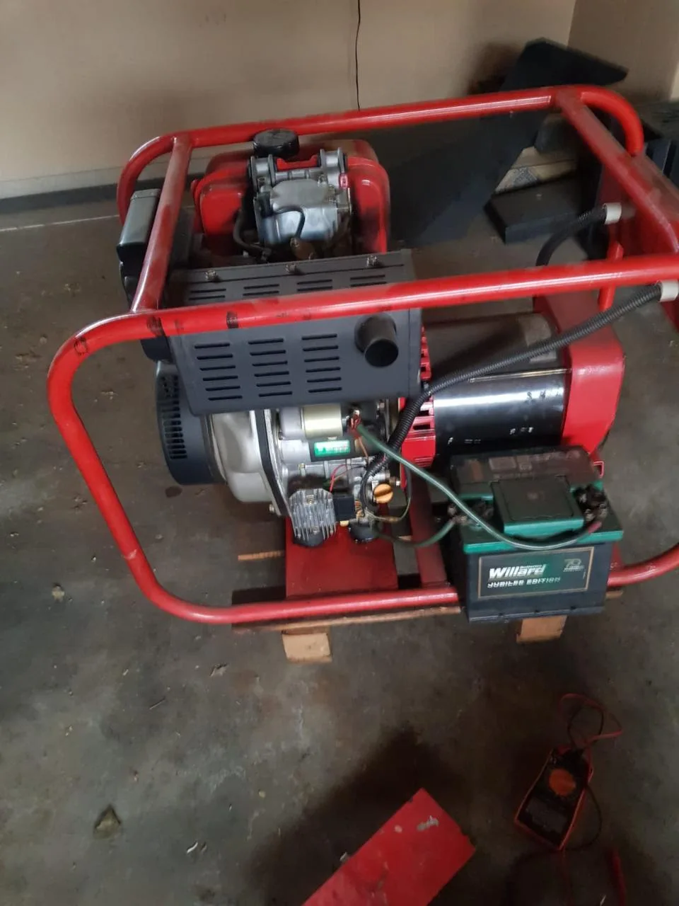 Red-framed open air-cooled diesel generator with electric start and a Willard battery on a garage floor, multimeter nearby