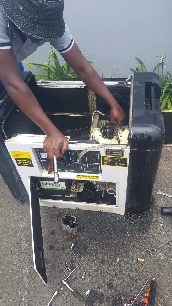 One person in a bucket hat working inside a stripped silent diesel generator, tools and parts on the ground