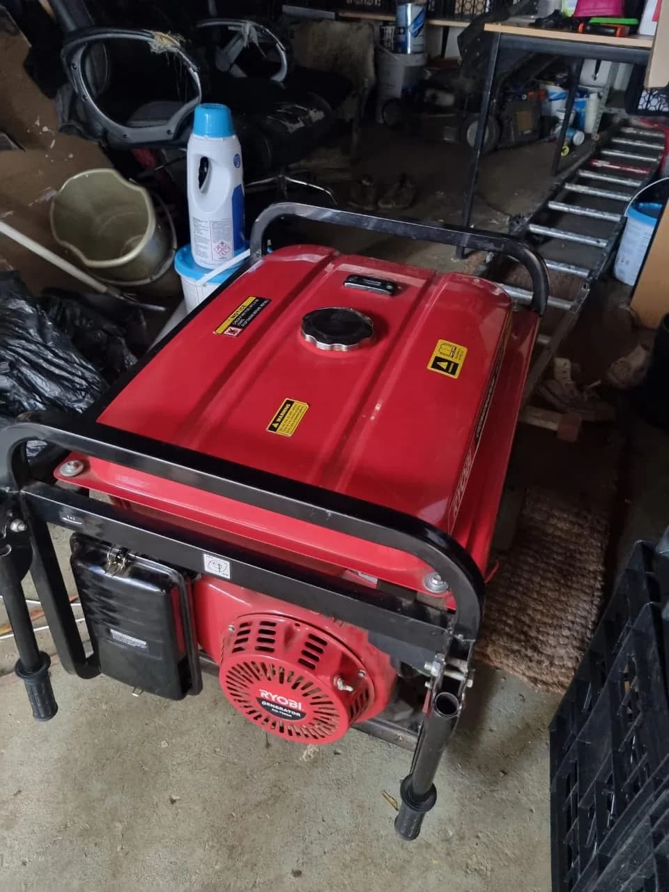 Red Ryobi portable generator with a recoil starter in a cluttered garage beside a bucket and shelving