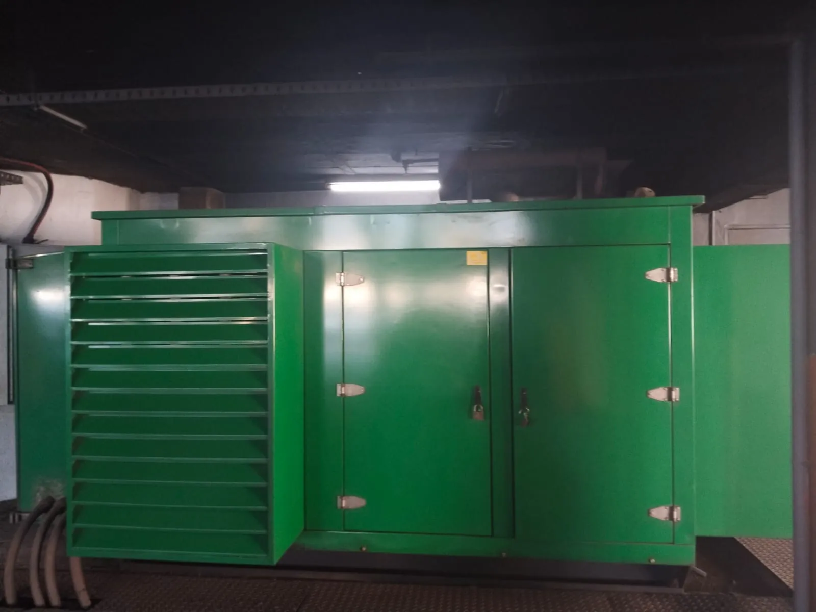 Green canopied diesel generator in a basement plant room under fluorescent lighting