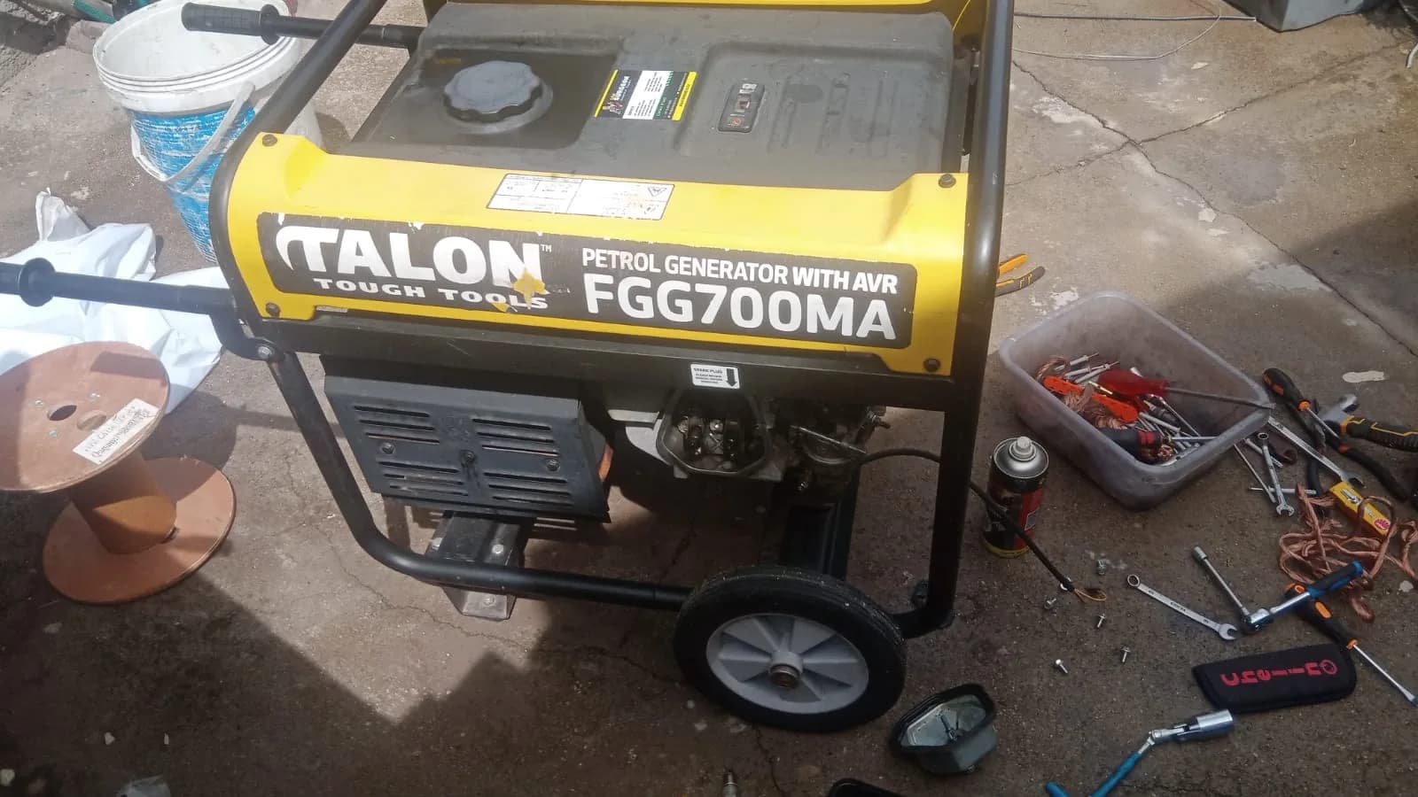 Yellow Talon FGG700MA petrol generator on concrete, with a tray of spanners, screwdrivers and parts beside it